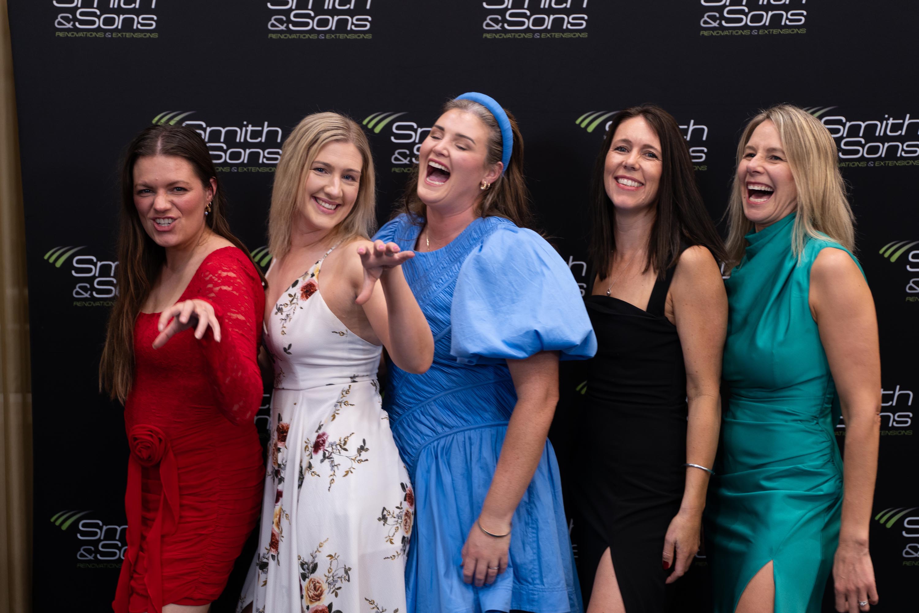 Five women laughing together in front of the branded Smith & Sons backdrop